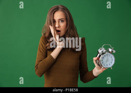 Image de jeune femme choquée isolés sur fond vert holding réveil. Banque D'Images