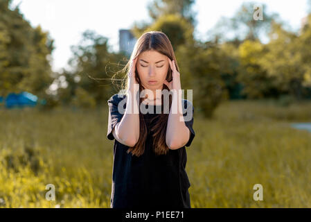 Fille brunette à l'air frais. Détient ses mains autour de sa tête. Des maux de tête. Essaie de se concentrer. La migraine et la douleur grave. Dans l'été dans le parc habillé en coagulation occasionnels. Banque D'Images