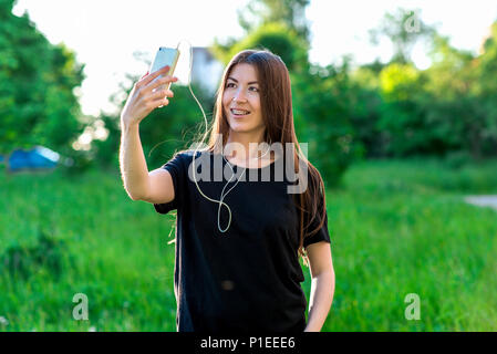 Fille asiatique brune en été dans le parc. Happy smiling dents appareil dentaire. Tient le téléphone dans le téléphone à l'écoute de la musique sur le casque. Réalise des vidéos. Il parle à travers les réseaux sociaux. Banque D'Images