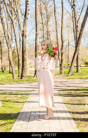 Femme parfaite en rose soyeux imperméable et boots holding bunch of flowers, mode extérieur portrait Banque D'Images