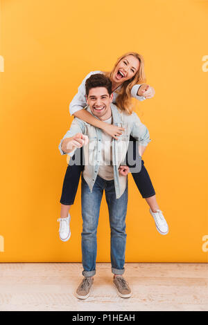 Photo de pleine longueur smiling couple having fun et montrer du doigt alors que vous man piggybacking woman isolated sur fond jaune Banque D'Images