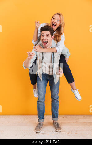 Longueur totale de l'image couple having fun et montrer du doigt copyspace tandis que man piggybacking femme joyeuse sur fond jaune isolé Banque D'Images