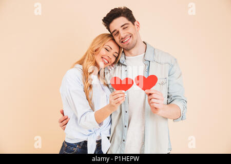 Image of man and woman in love portant des vêtements en jean serrant ensemble avec les yeux fermés et la tenue de deux coeurs en papier rouge plus isolés b beige Banque D'Images