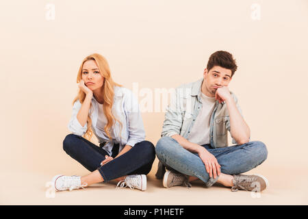 Insatisfait de l'homme et la femme assise sur le sol avec les jambes croisées et soutenir leurs têtes après lutte ou querelle isolated over beige background Banque D'Images