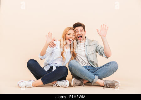 Photo de heureux deux amis l'homme et la femme dans des vêtements décontractés assis sur le sol avec les jambes croisées et l'accueil avec des signes avec la main isolated over beige Banque D'Images