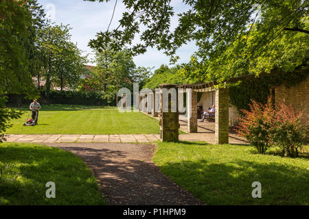 Un jardinier qui tond la pelouse à Malmesbury Abbey Gardens, alors qu'un visiteur se trouve dans le soleil et des silences, Wiltshire, UK Banque D'Images