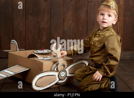 Les enfants garçon sont habillés en soldat en uniforme militaire retro réparer un avion en carton fort, fond en bois foncé, style retro Banque D'Images