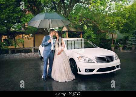 Beau couple de jeunes mariés sur fond de voiture blanche avec parapluie Banque D'Images