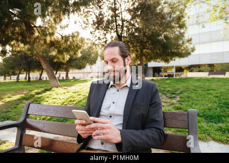 Photo de l'homme barbu 30s dans l'usure formelle holding et la saisie sur téléphone cellulaire tout en vous reposant à city park Banque D'Images