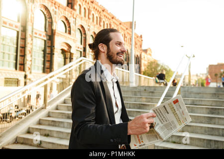 Photo gros plan de l'homme souriant 30s en costume officiel walking in city street et lisant le journal d'économie Banque D'Images