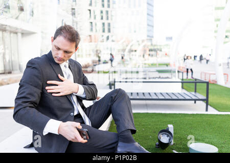 Jeune homme d'époussetage nettoyage off suit sitting on bench in urban green Park à la recherche vers le bas avec cravate sur pause interview Banque D'Images