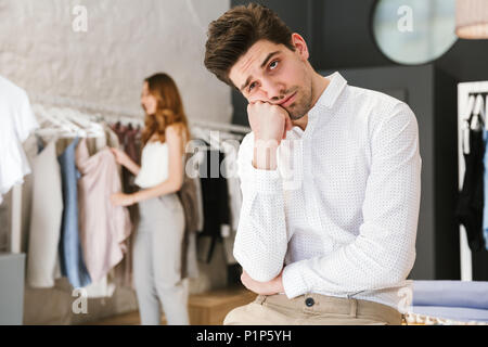 Homme fatigué assis et à la recherche à l'appareil photo pendant que sa petite amie faire les boutiques de vêtements sur un arrière-plan Banque D'Images