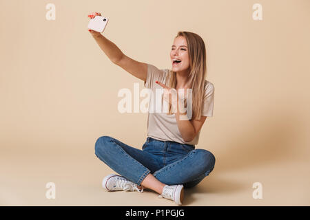 Blonde woman gesturing index au sens de l'appareil photo et vous prendre photo sur selfies cell phone while sitting in lotus pose plus isolés Banque D'Images