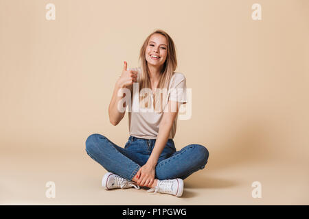 Cheerful blonde woman sitting on floor in lotus pose et showing thumb up at camera isolated over beige background Banque D'Images