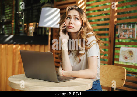 Portrait of a sad young girl talking on mobile phone alors qu'il était assis avec un ordinateur portable dans un café en plein air Banque D'Images