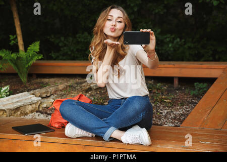 Belle jeune fille prenant tout en selfies assis sur un banc à l'extérieur et l'envoi de kiss Banque D'Images