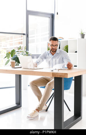 Longueur totale de droit d'affaires unshaved 30s en chemise blanche working in office sur les ordinateurs portables et à la recherche au presse-papiers avec les documents papier Banque D'Images