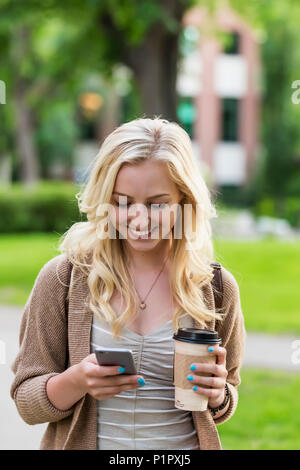 Une belle jeune femme avec de longs cheveux blonds tenant une tasse de café et des sms sur son téléphone intelligent en vous promenant dans un campus universitaire Banque D'Images