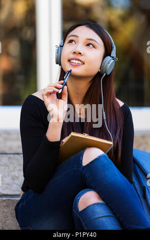 Une jeune étudiante chinoise se trouve sur les marches à écouter de la musique à l'aide d'un casque et regarde tout en rêvant et en tenant un stylo pour... Banque D'Images