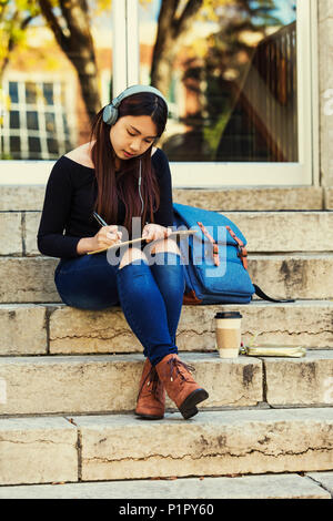 Un jeune étudiant d'université chinois est assis sur les marches d'écouter de la musique à l'aide d'écouteurs et écrit dans l'ordinateur portable ; Edmonton, Alberta, Canada Banque D'Images