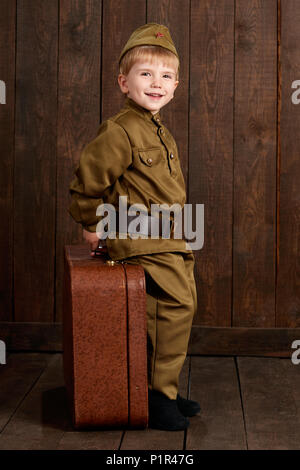 Les enfants garçon sont habillés en soldat en uniforme militaire rétro avec vieille valise, fond en bois foncé, style retro Banque D'Images