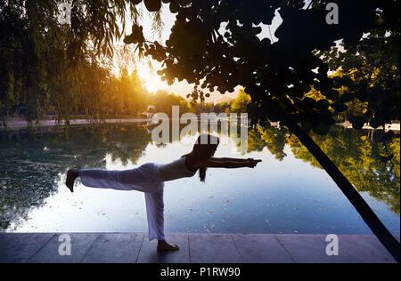 Belle fille asiatique en costume blanc faisant du yoga près du lac, dans le parc, au lever du soleil Banque D'Images