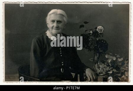 Allemagne - circa 1930 : Vintage photo montre femme âgée est assise sur le fauteuil de la période dans un studio de photographie. Sur la table sont des fleurs. Retro noir et blanc photographie studio avec effet sépia. Banque D'Images