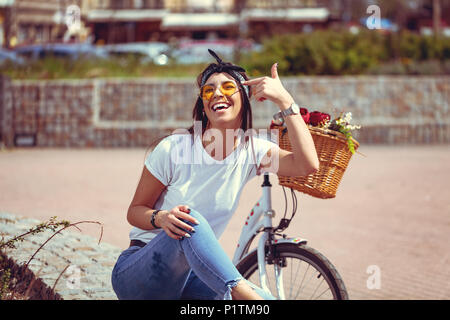 Jolie jeune femme est heureux de s'amuser et profiter de mettre un doigt sur son front, dans une journée ensoleillée d'été, à côté du vélo avec panier de fleurs. Banque D'Images
