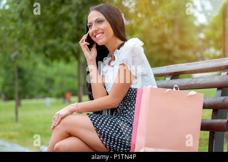 Cheerful girl après shopping avec de nombreux sacs shopping assis sur un banc dans le parc et de parler au téléphone intelligent. Banque D'Images