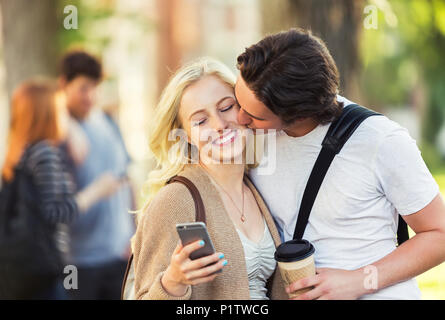 Un jeune homme embrasse une jeune femme sur la joue alors qu'elle est contrôle de médias sociaux sur un téléphone intelligent sur un campus universitaire ; Edmonton, Alberta, Canada Banque D'Images