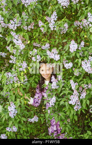 Young beautiful smiling girl se cachant dans des buissons de lilas en fleurs Banque D'Images