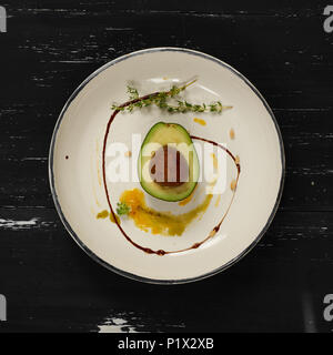 L'avocat coupé en deux demi-parties décorées avec le thym, la sauce jaune et les écrous de cèdre couché sur plaque ronde plate. Restaurant délicieux repas vegan pour les matières foo Banque D'Images