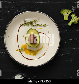 L'avocat coupé en deux demi-parties décorées avec le thym, la sauce jaune et les écrous de cèdre couché sur plaque ronde plate. Restaurant délicieux repas vegan pour les matières foo Banque D'Images