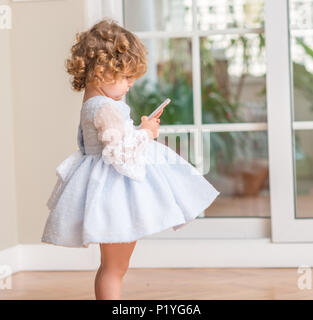 Belle blonde dans une robe enfant debout jouant avec smartphone à la maison. Banque D'Images