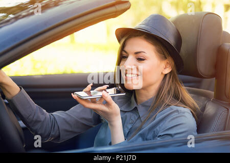 Heureux femme assise à l'intérieur voiture décapotable en utilisant un téléphone intelligent fonction reconnaissance vocale à la ligne à l'extérieur, sur un arrière-plan lumineux park Banque D'Images