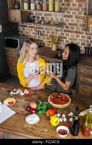 Souriante jeune femme multiethnique tintement des verres à vin pendant la cuisson de la pizza Banque D'Images