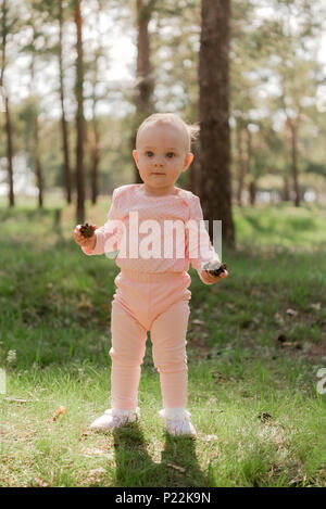 Le bébé tient deux cônes dans la forêt d'épinettes contre l'herbe verte dans un costume rose et a l'air passé l'appareil photo Banque D'Images