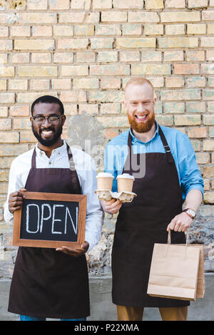 Les jeunes baristas multiethnique avec inscription ouvrir, sacs en papier et de tasses à café jetables smiling at camera Banque D'Images