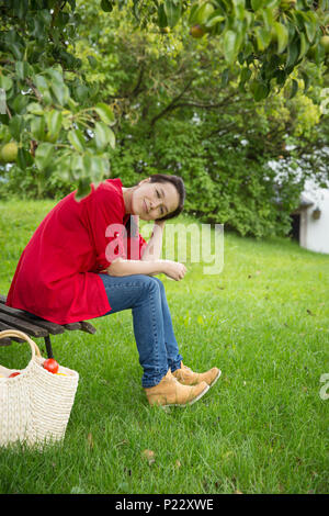 Belle jeune femme assise sur un banc en bois sous un poirier et souriant vers un appareil photo Banque D'Images