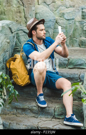 Beau jeune homme taking photo with smartphone tout en étant assis sur un escalier en pierre Banque D'Images