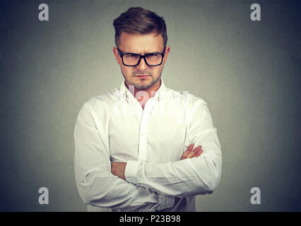 Jeune homme sérieux dans les verres tenant les mains croisées et à la recherche à l'appareil photo en n'aiment pas sur fond gris. Banque D'Images