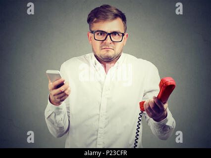 Young man holding old-fashioned red phone et smartphone moderne looking at camera dans la perplexité. Banque D'Images