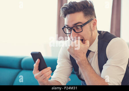 Jeune homme dans l'appartement regardant des smartphone et de mordre dans l'expression de la peur le poing. Banque D'Images