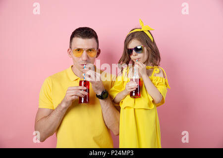 Vue de la récolte de jeune homme et enfant mignon dans des tenues jaunes holding red boissons avec des pailles à la caméra et au sourire sur fond rose Banque D'Images