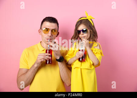 Vue de la récolte de jeune homme et enfant mignon dans des tenues jaunes holding red boissons avec des pailles à la caméra et au sourire sur fond rose Banque D'Images