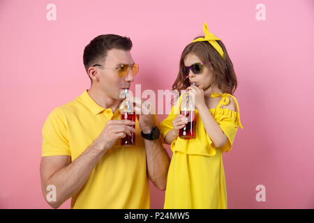 Vue de la récolte de jeune homme et enfant mignon dans des tenues jaunes holding red boissons avec des pailles à l'un l'autre et souriant sur fond rose Banque D'Images