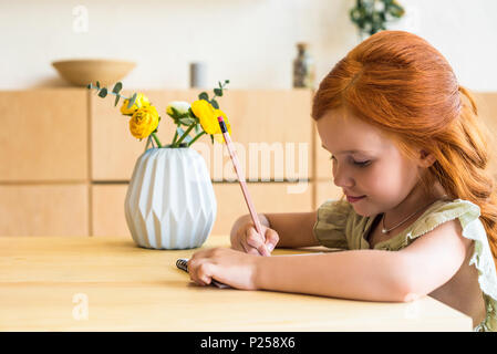 Vue de côté de fille rousse mignon le dessin avec un crayon tout en étant assis à table Banque D'Images