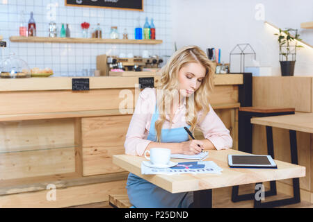 Jeune femme sérieuse cafe owner taking notes while sitting at table Banque D'Images