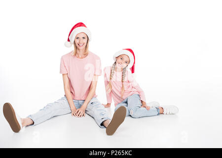 Happy mother and daughter wearing santa hats and sitting on floor, isolated on white Banque D'Images