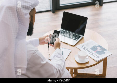 Homme d'affaires musulman en utilisant smartphone pendant que sitting on couch with laptop on table Banque D'Images